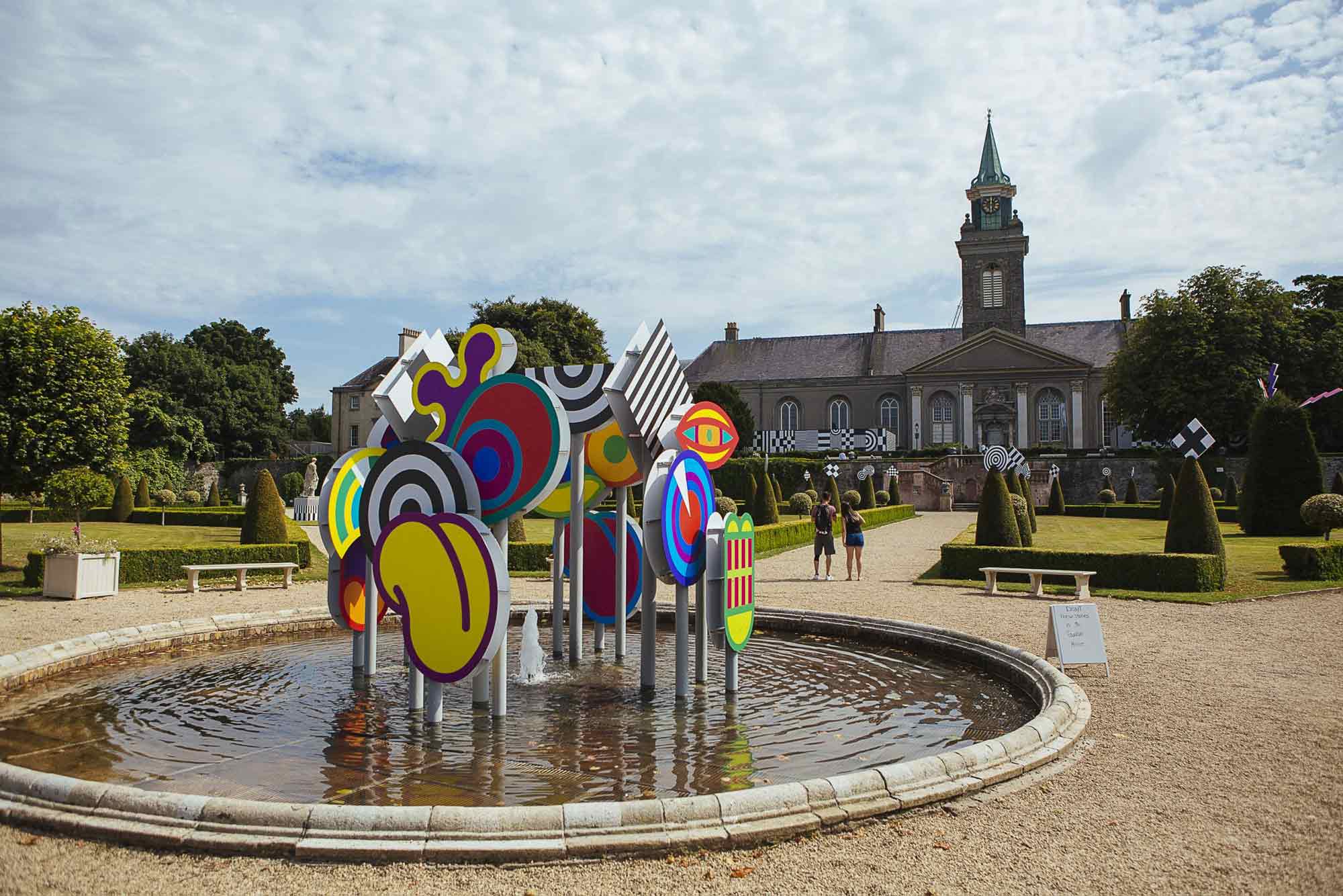 Exterior installation photograph of a fountain in a formal garden, an installation of colourful shapes is in the centre of the fountain.